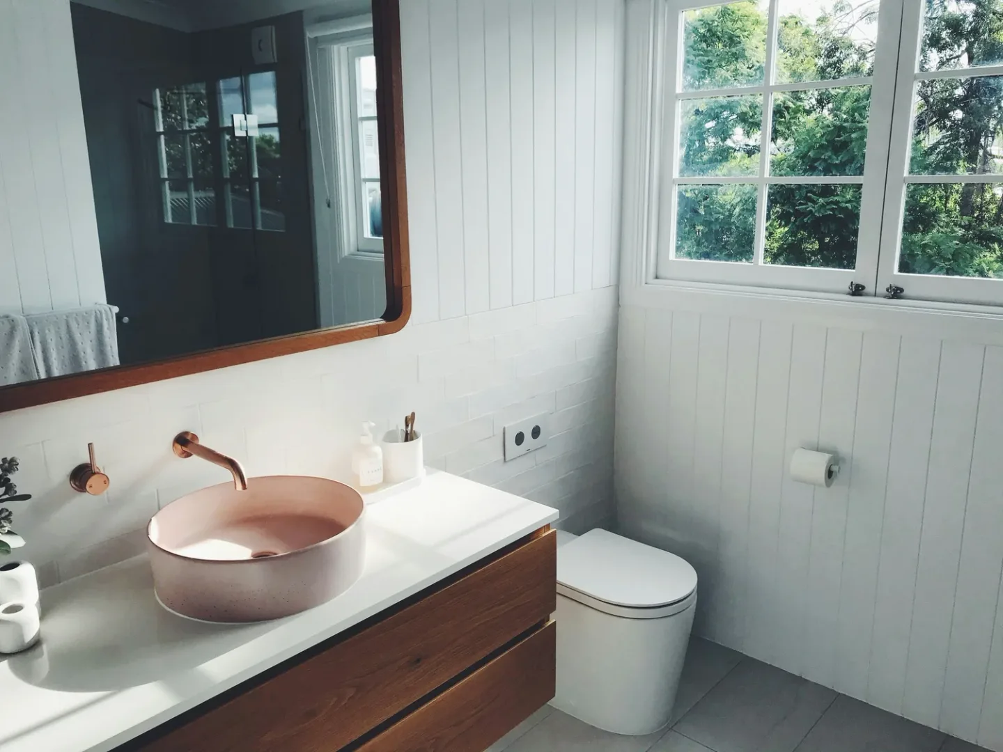 Modern bathroom with a light pink round sink on a white countertop, wooden vanity, white panel walls, a large window with trees outside, a mirror with a wooden frame, and a toilet.